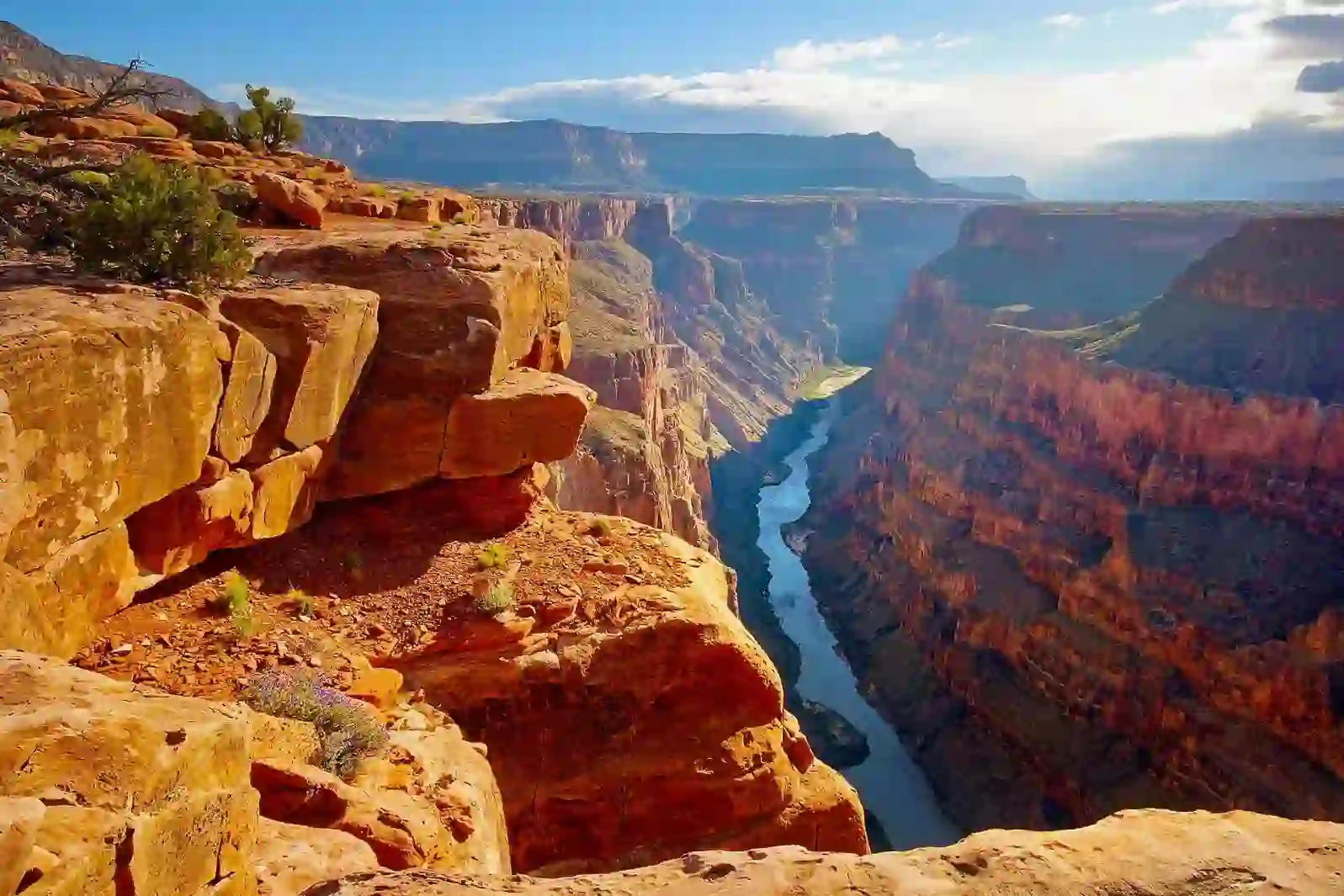 A breathtaking view of the Grand Canyon with its layered red rock cliffs and the Colorado River winding through the valley under a partly cloudy sky