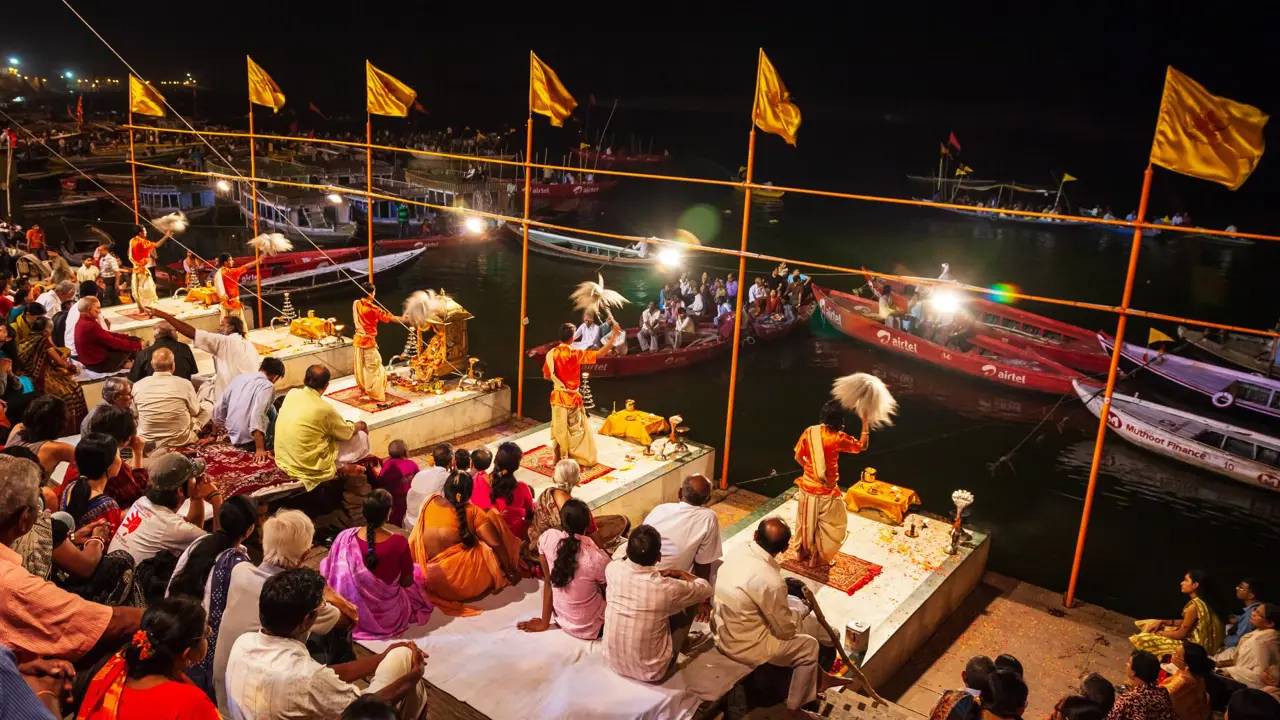 Priests dressed in traditional saffron robes performing the Ganga Aarti ceremony on the ghats of Varanasi at night, surrounded by devotees and illuminated by rows of lights along the riverbank