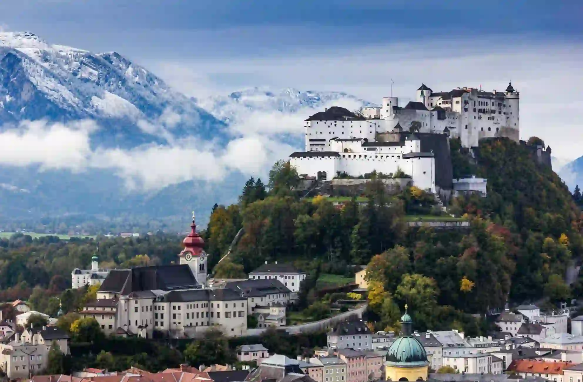 View of the Hohensalzburg Fortress, a white castle with black roofs on top of a grassy hill. At the bottom of the hill is a white large building with a red turret. Behind these is a large mountain with snow on and clouds in front of it.