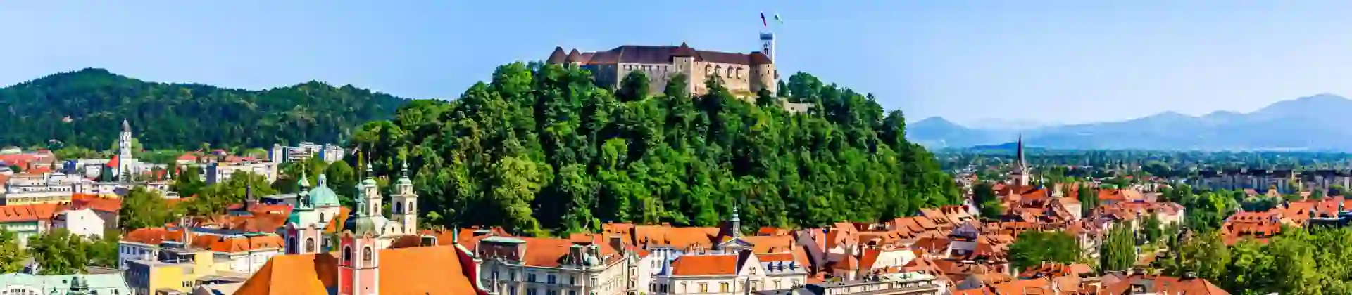 High angle shot of a town, showing lots of buildings with orange roofs and a hill in the centre covered in trees, with a large stone building on top. Mountains in the distance on the right and a mountain covered in trees on the left.