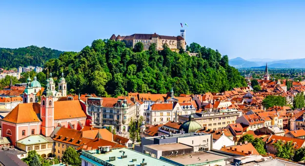 High angle shot of a town, showing lots of buildings with orange roofs and a hill in the centre covered in trees, with a large stone building on top. Mountains in the distance on the right and a mountain covered in trees on the left.