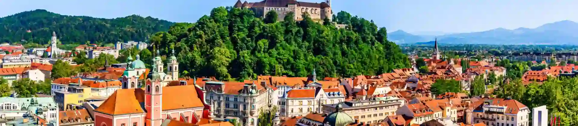 High angle shot of a town, showing lots of buildings with orange roofs and a hill in the centre covered in trees, with a large stone building on top. Mountains in the distance on the right and a mountain covered in trees on the left.