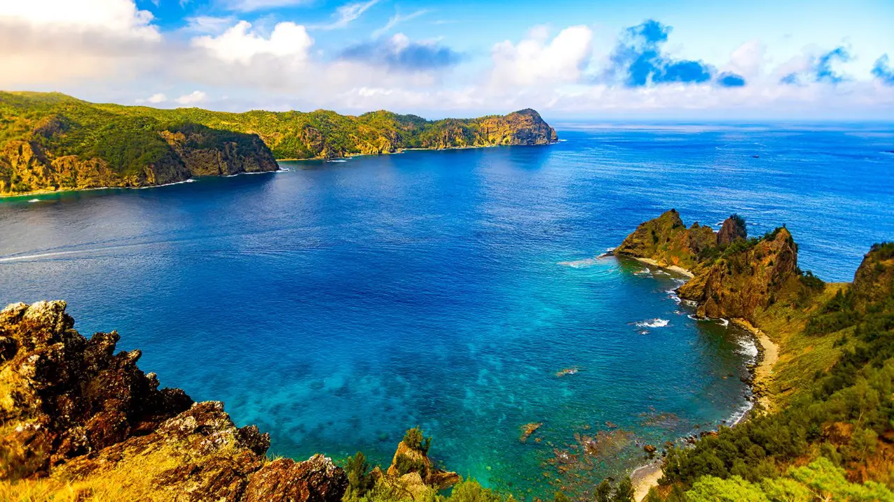 A stunning coastal view from Nagasaki Observatory on Chichijima Island, with clear blue waters, rugged cliffs, and lush green hills under a vibrant sky