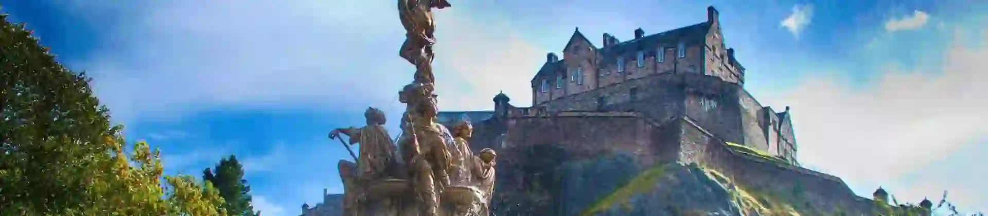 A view of Edinburgh Castle perched high on a rocky hill in the background, with a decorative fountain in the foreground below, surrounded by greenery