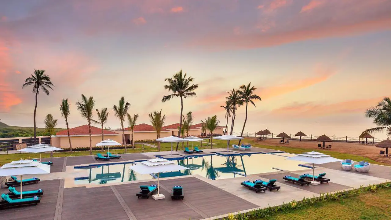 The outdoor pool at Welcomhotel by ITC Hotels, Mahabalipuram, glows at sunset, surrounded by palm trees and reflecting the warm orange sky
