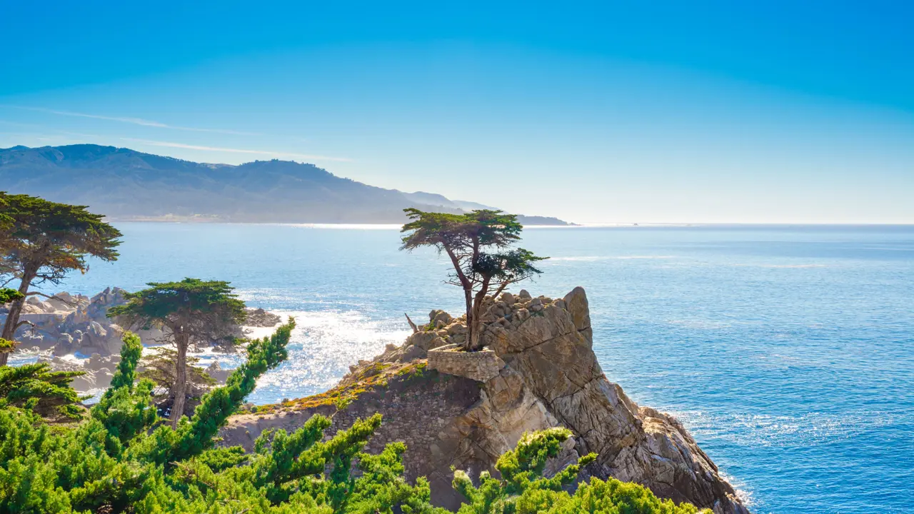 Coastal view of Pebble Beach, California, with rugged shoreline, rocky outcrops, and waves crashing against the coast under a clear blue sky