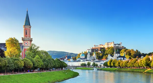 View of a river winding to the left, with grass and trees on either side, and a red church with a tall, slim tower with a blue turret. On the left of the river multiple large buildings can be seen, with numerous turquoise turrets pointing out, and the fortress on the top of the hill, all under a blue sky