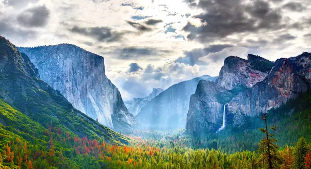 Yosemite National Park in California with towering granite cliffs, green forested areas, and cloudy skies with sun rays breaking through