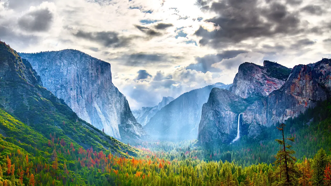 Yosemite National Park in California with towering granite cliffs, green forested areas, and cloudy skies with sun rays breaking through