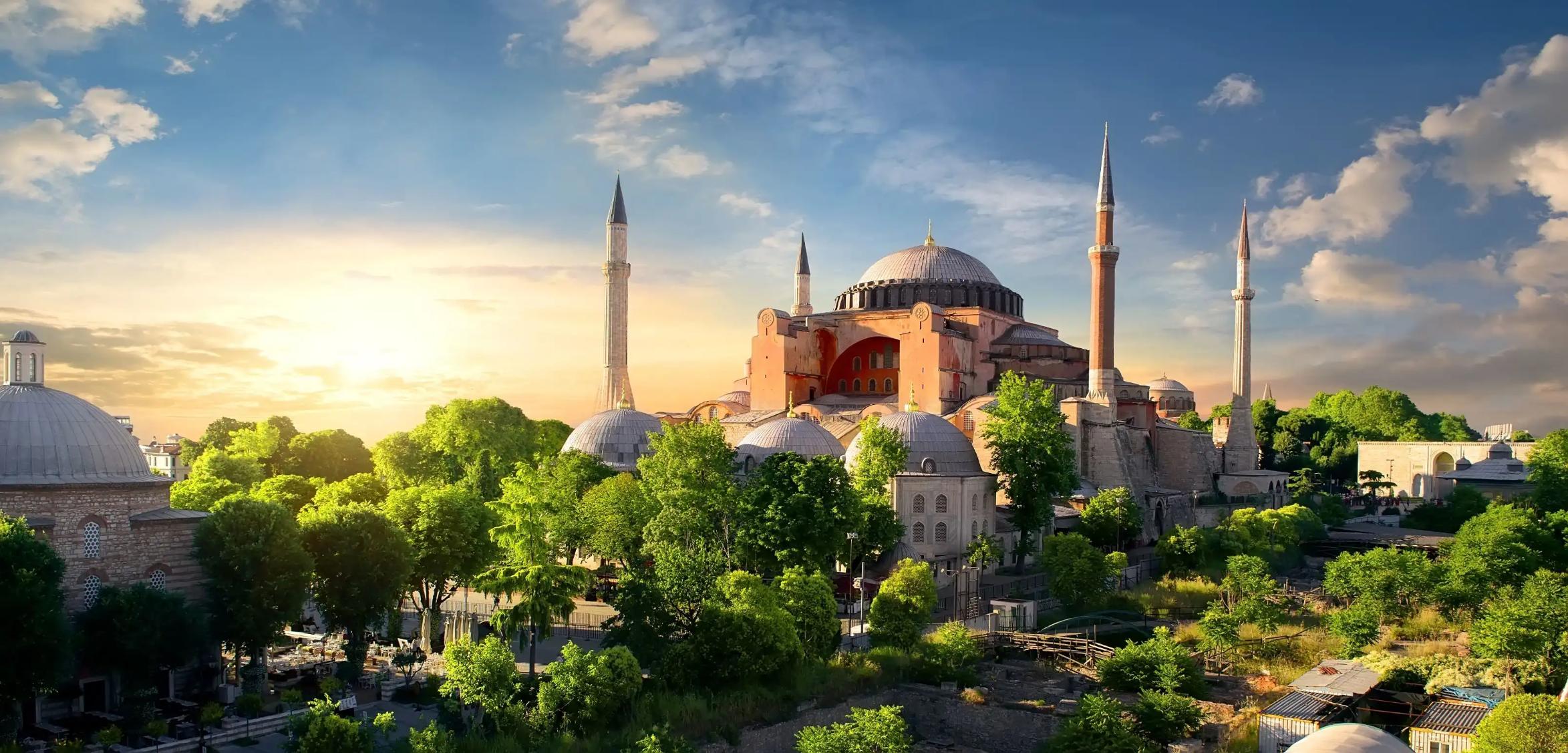 Hagia Sophia with its domes and minarets at sunset, surrounded by green trees in Istanbul