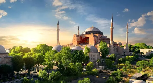 Hagia Sophia with its domes and minarets at sunset, surrounded by green trees in Istanbul