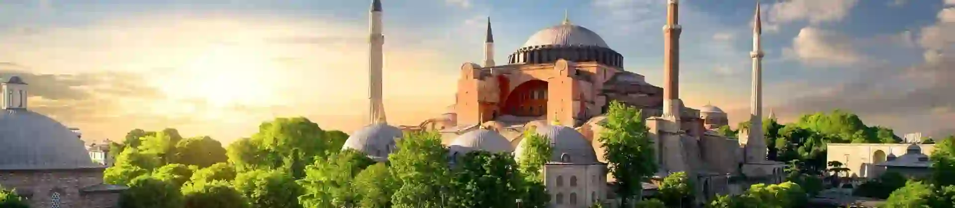 Hagia Sophia with its domes and minarets at sunset, surrounded by green trees in Istanbul