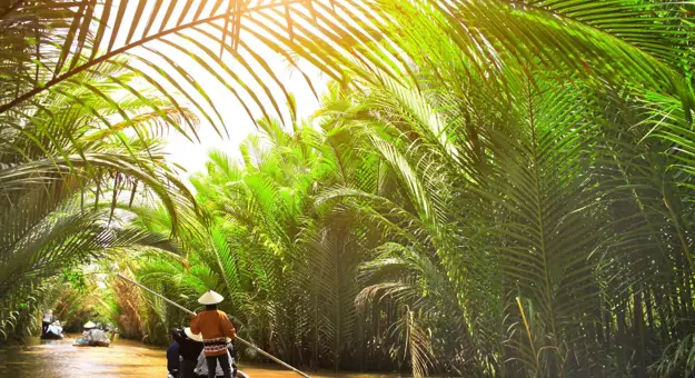 Small wooden boats glide through a palm-lined canal off the Mekong River in Thailand, with a person in a conical hat steering under filtered sunlight
