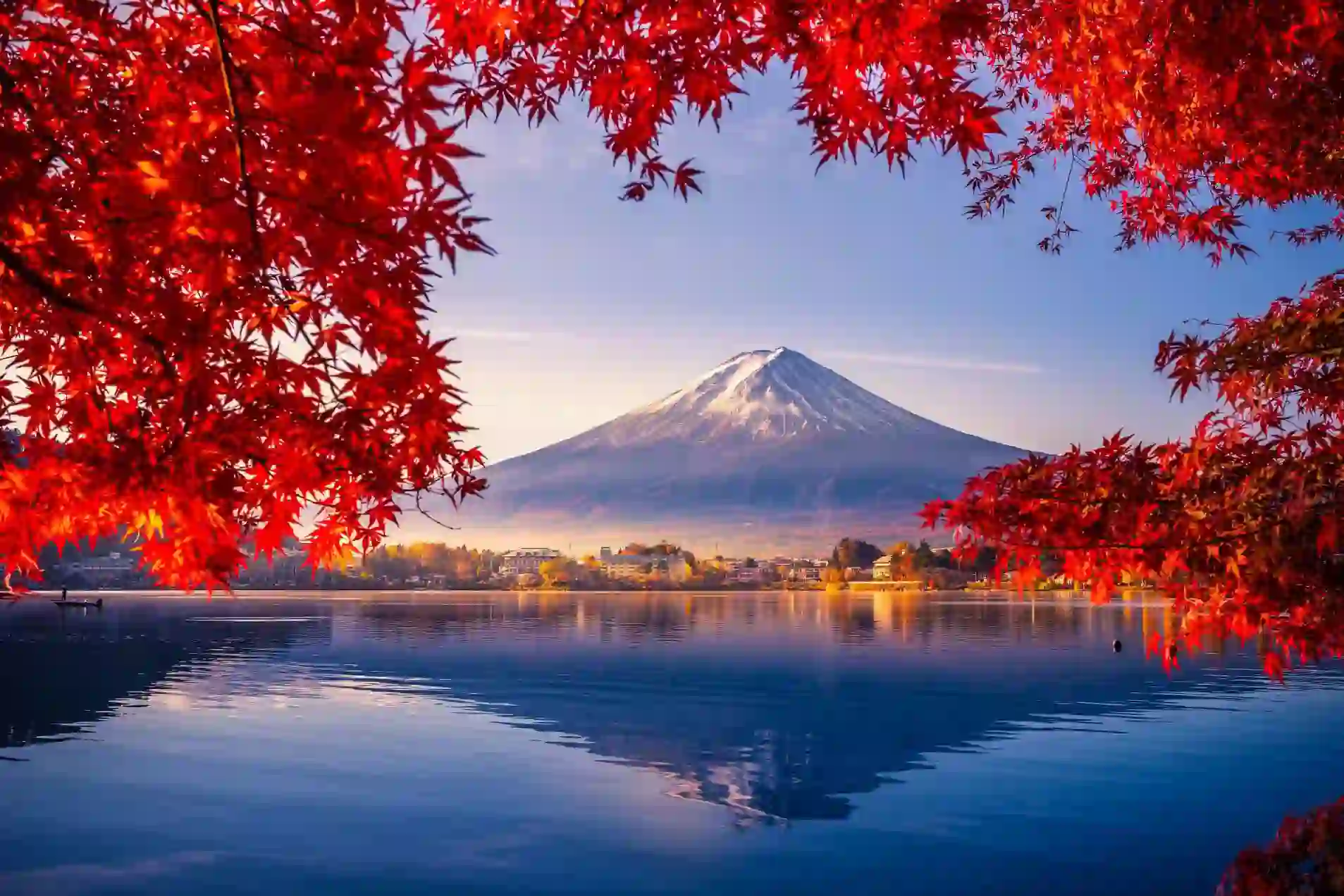 Mount Fuji reflected in Lake Kawaguchi, framed by vivid red autumn leaves, with a sunlit lakeside town between the water and the mountain