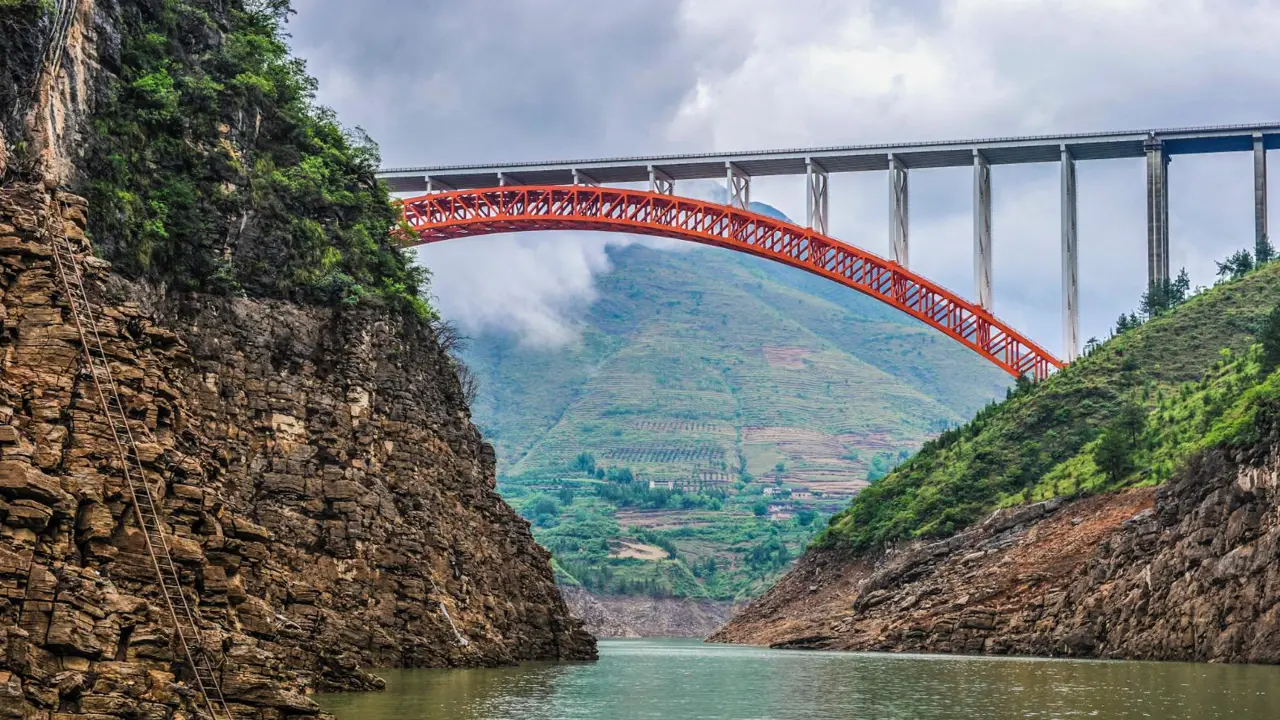 A red arch bridge spanning between steep cliffs above the Yangtze River, surrounded by green mountains and mist, China