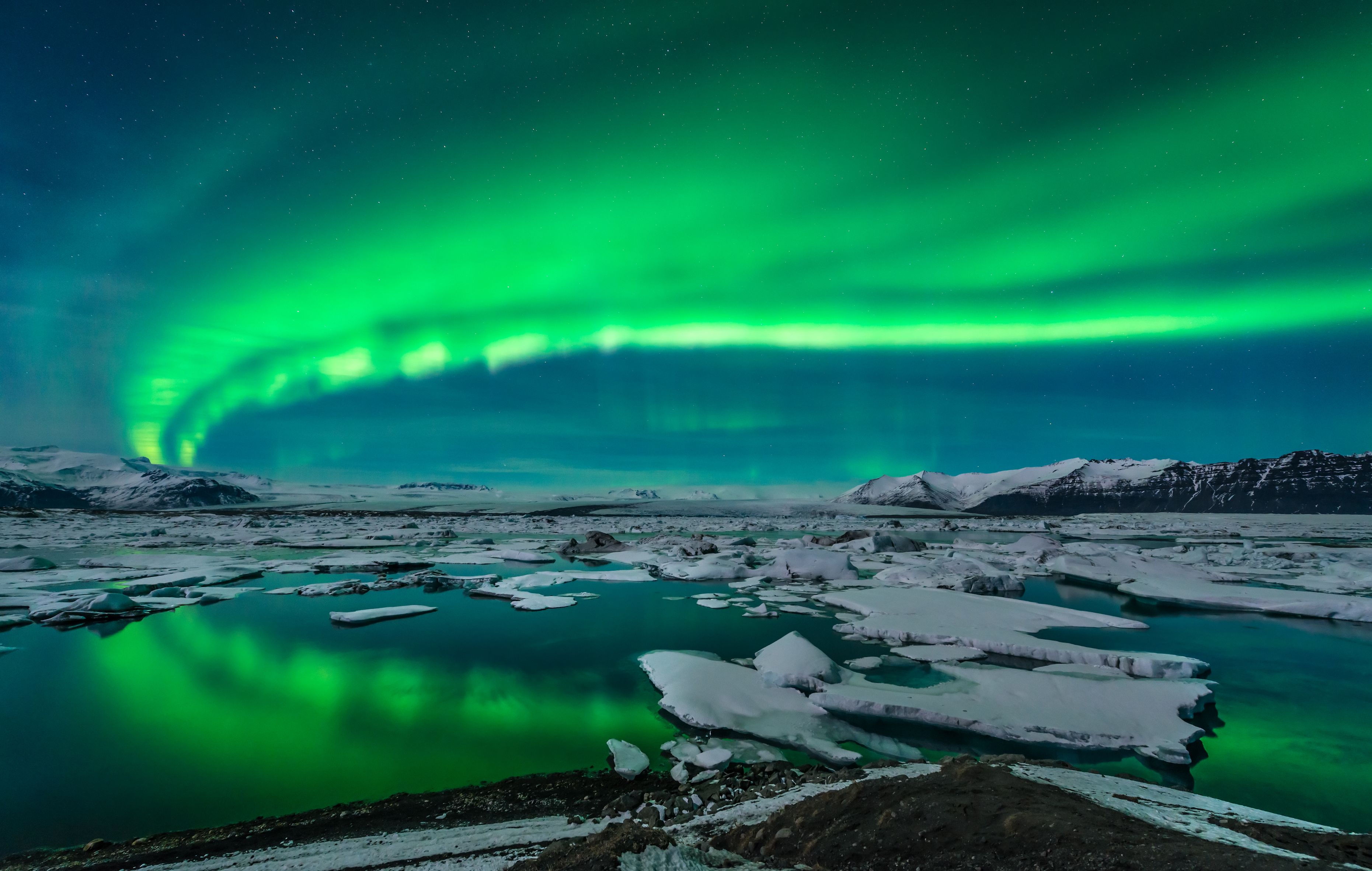Green northern lights curving from the right back to the left. Below, a lake with ice blocks
