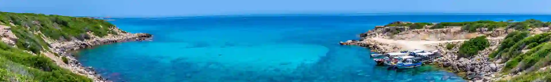 View of the sea from land, the edge being a circular shape with some boats docked to the right. The land has some grass and the sky is blue.