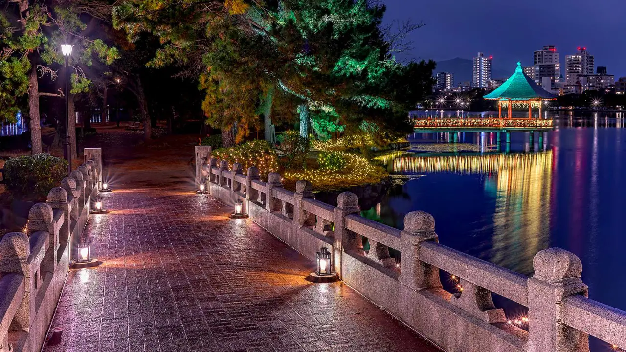 A panoramic cityscape of Fukuoka at night, showing modern buildings, a river, and green urban spaces under a partly cloudy sky