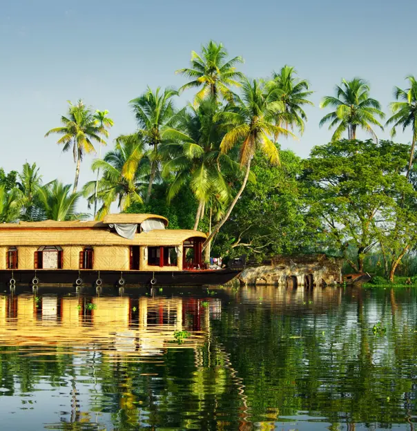 A light brown wicker houseboat on calm water in Kerala, with palm trees on the banks and clear blue skies overhead