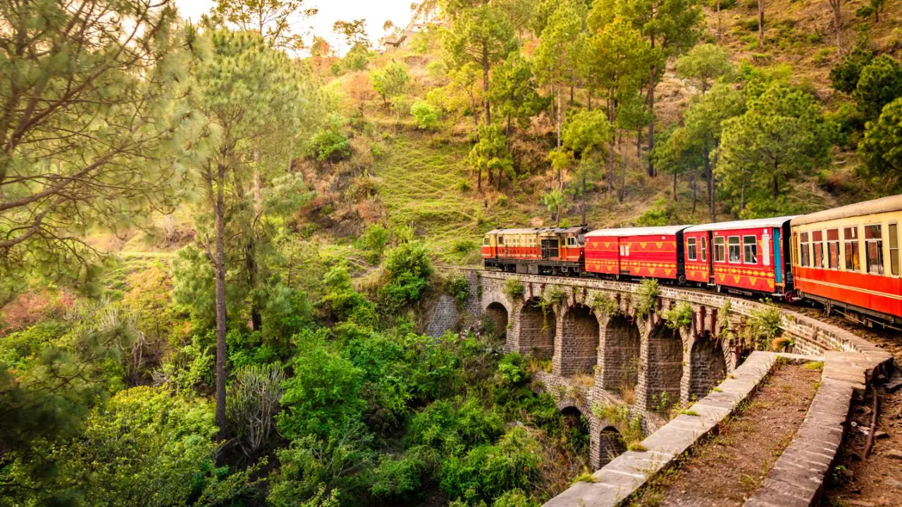 The heritage toy train crossing a railway bridge on the Kalka–Shimla route, surrounded by lush green hills and trees