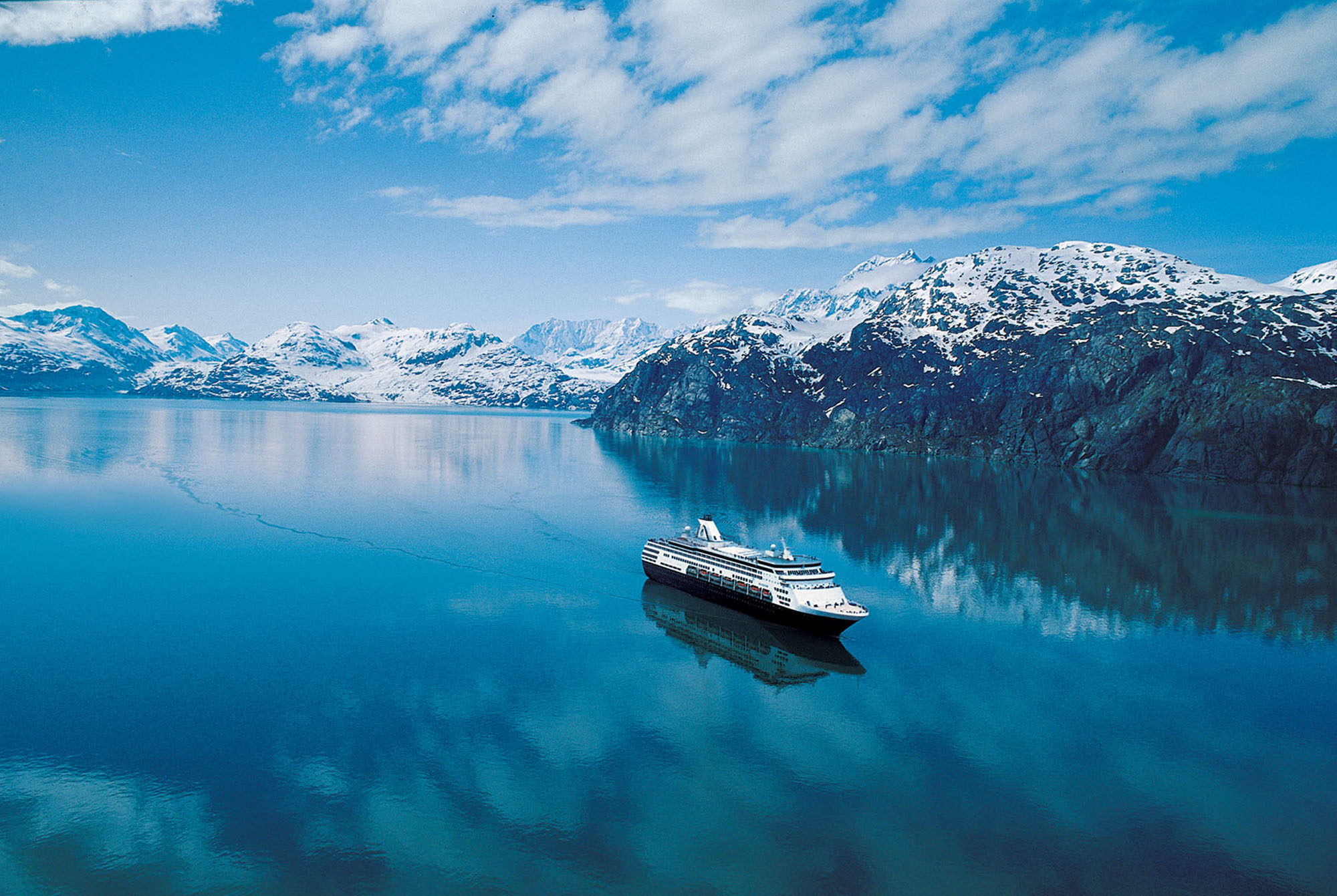 A Holland America Line cruise ship sailing through the clear blue waters of the Inside Passage near Alaska, with snow-capped mountains in the distance