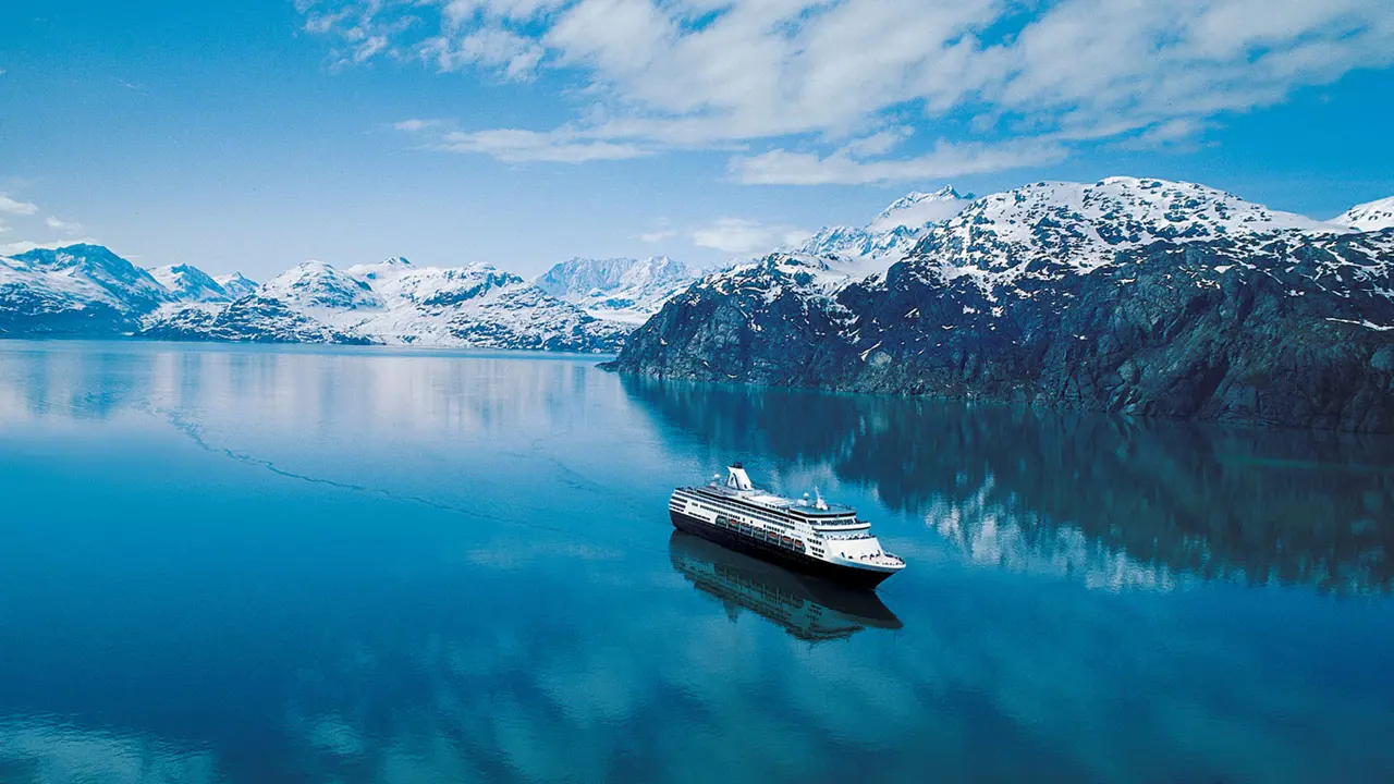 A Holland America Line cruise ship sailing through the clear blue waters of the Inside Passage near Alaska, with snow-capped mountains in the distance