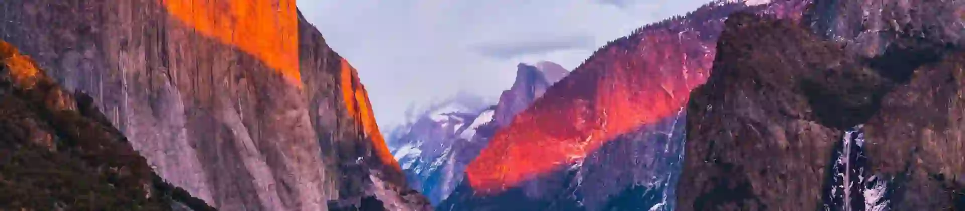 A dramatic view of Yosemite’s El Capitan and surrounding mountains bathed in warm, glowing sunset light under a cloudy sky