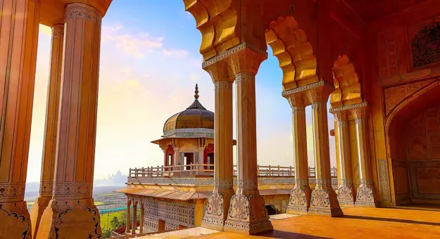 A view through ornate sandstone arches at Agra Fort, overlooking a domed pavilion and distant landscape in warm evening light