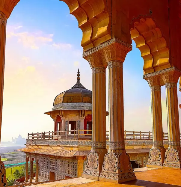 A view through ornate sandstone arches at Agra Fort, overlooking a domed pavilion and distant landscape in warm evening light