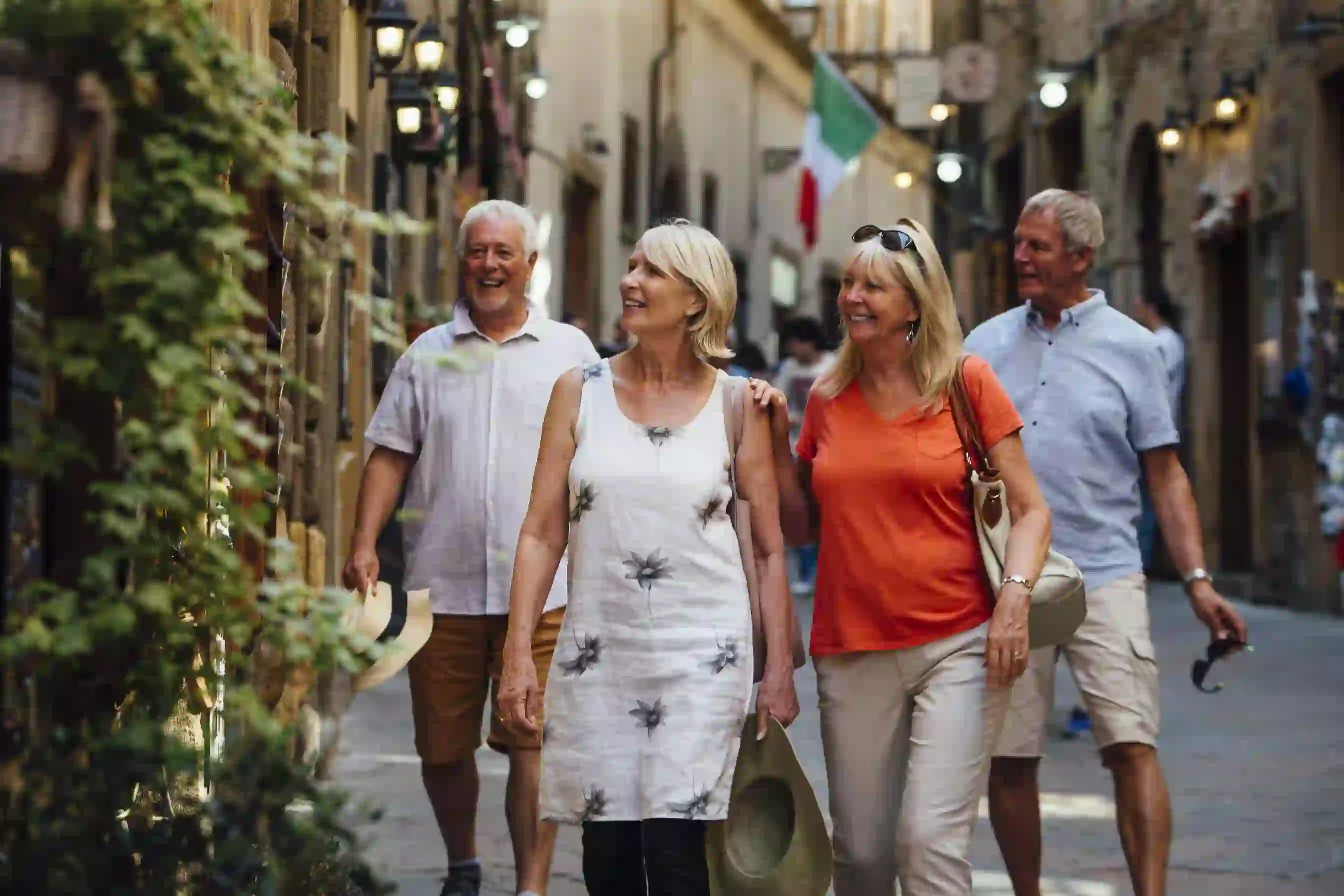 Smiling elderly friends stroll through a charming Italian street with historic buildings and an Italian flag in the background