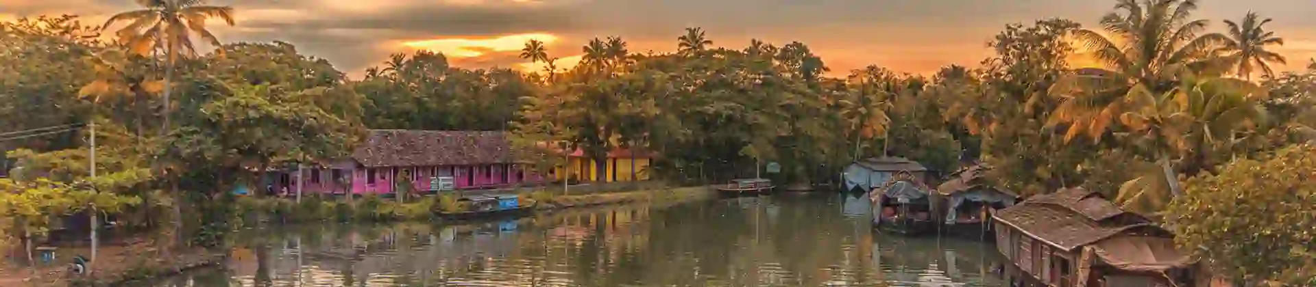 Two people in a canal boat on the Kerala Backwaters at sunset, with a pink house and palm trees lining the banks, and warm light reflecting on the calm water