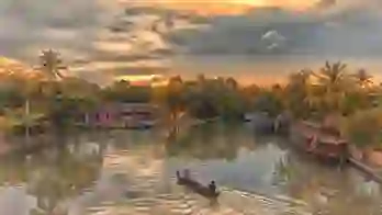 Two people in a canal boat on the Kerala Backwaters at sunset, with a pink house and palm trees lining the banks, and warm light reflecting on the calm water