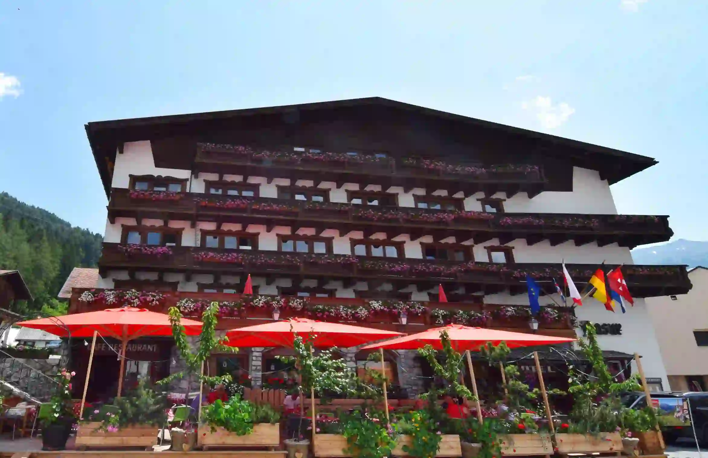 A traditional alpine-style hotel in Austria with wooden balconies decorated with pink and white flowers. Red parasols shade an outdoor café area which is surrounded by greenery, and several international flags are displayed at the front