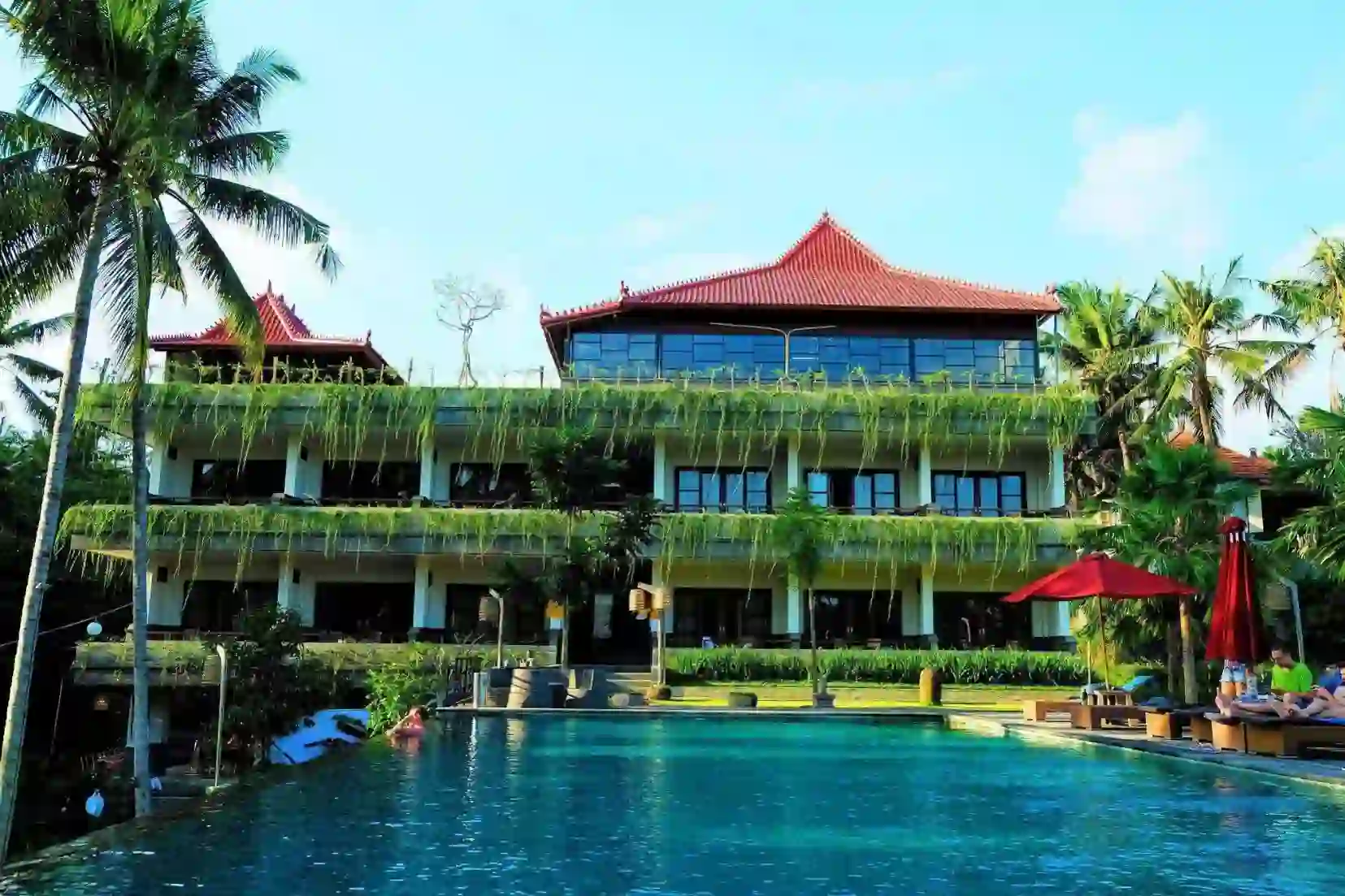 Exterior of Artini Dijiwa Hotel in Ubud, with balconies covered in greenery and a swimming pool in front, surrounded by palm trees
