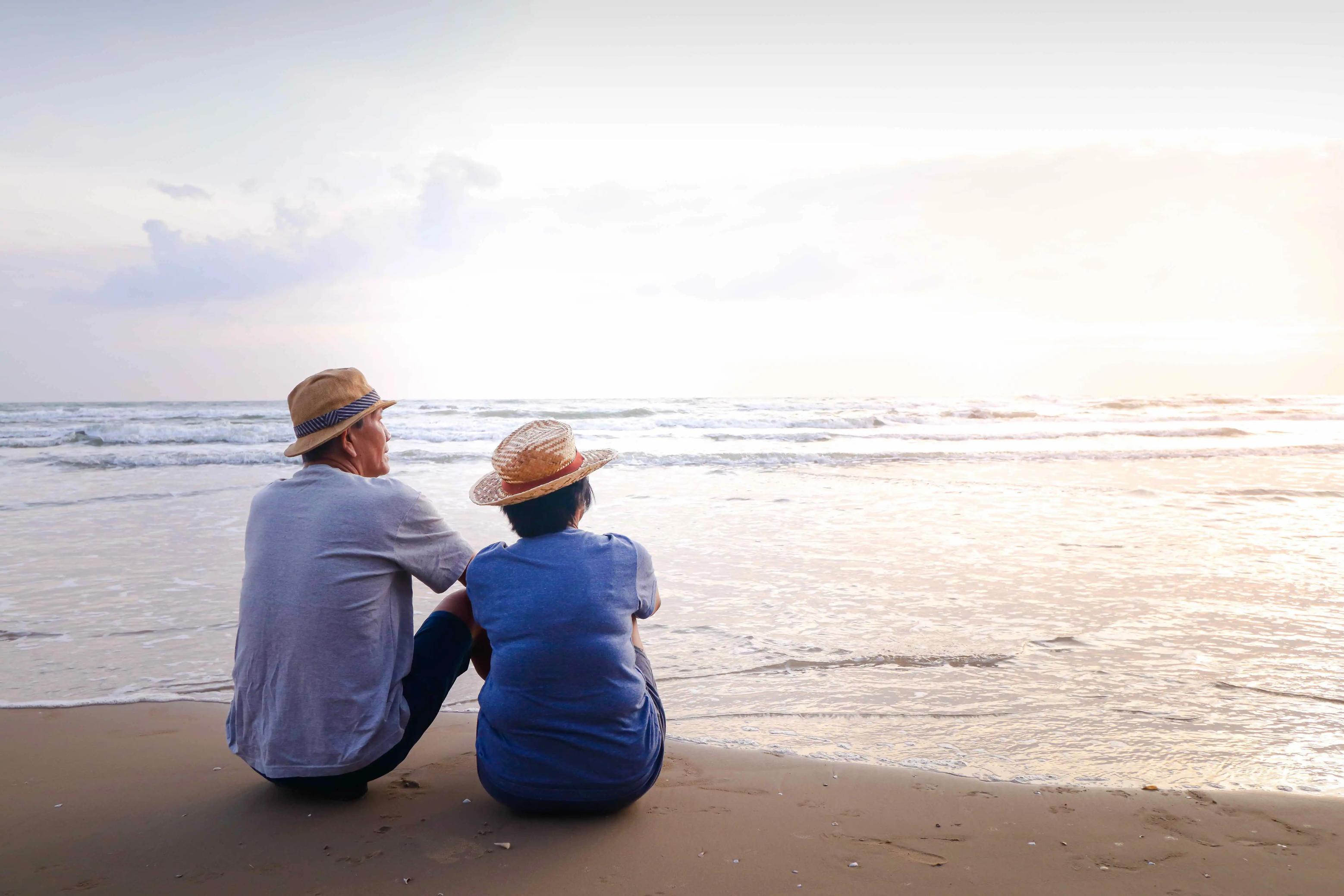 An elderly couple in straw hats sits side by side on a sandy beach, peacefully watching the calm ocean as the sun sets