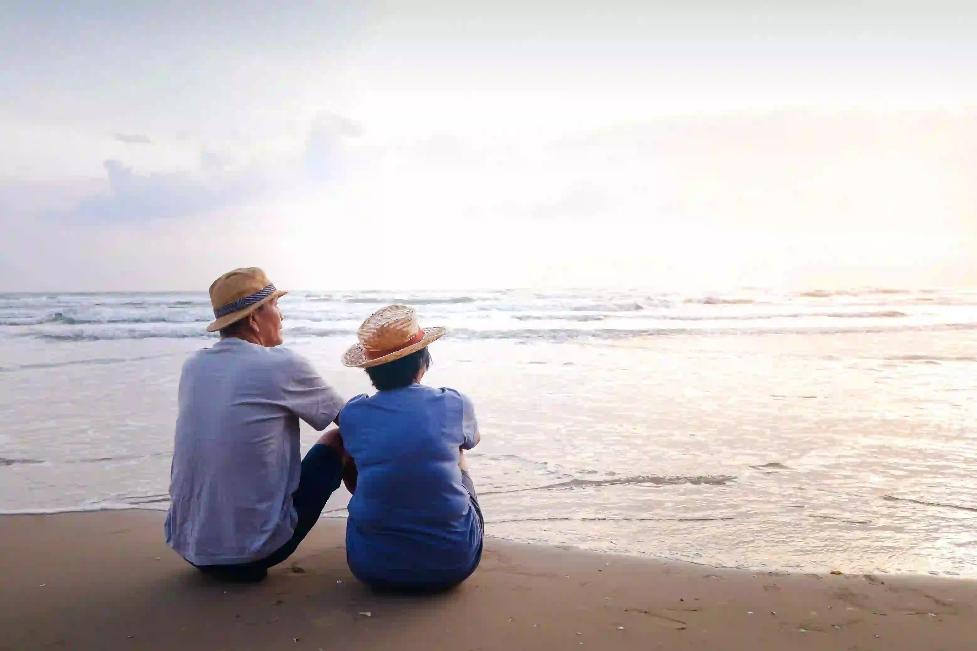 An elderly couple in straw hats sits side by side on a sandy beach, peacefully watching the calm ocean as the sun sets