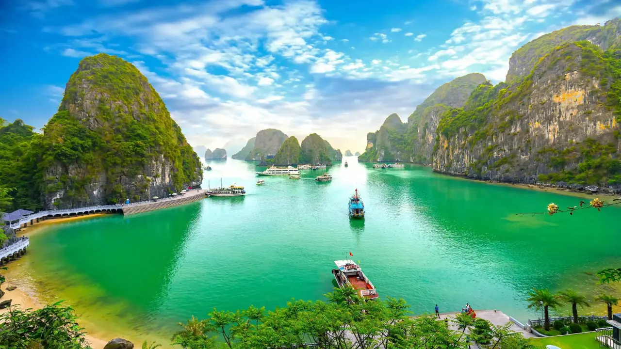 Panoramic view of Ha Long Bay in Vietnam, with emerald green waters dotted with traditional wooden boats, surrounded by towering limestone karsts and lush, forested islets under a partly cloudy sky