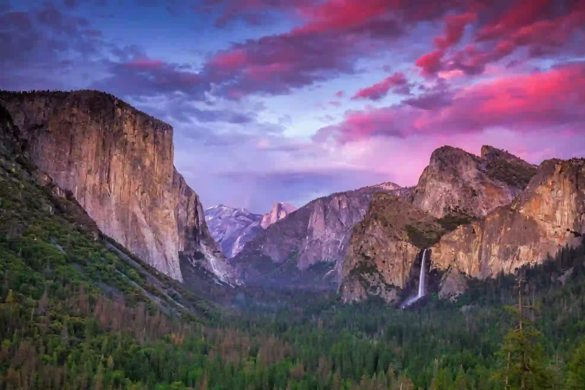 Breathtaking view of Yosemite National Park, California, featuring towering granite cliffs and lush green forests beneath a pink and blue sky at sunset