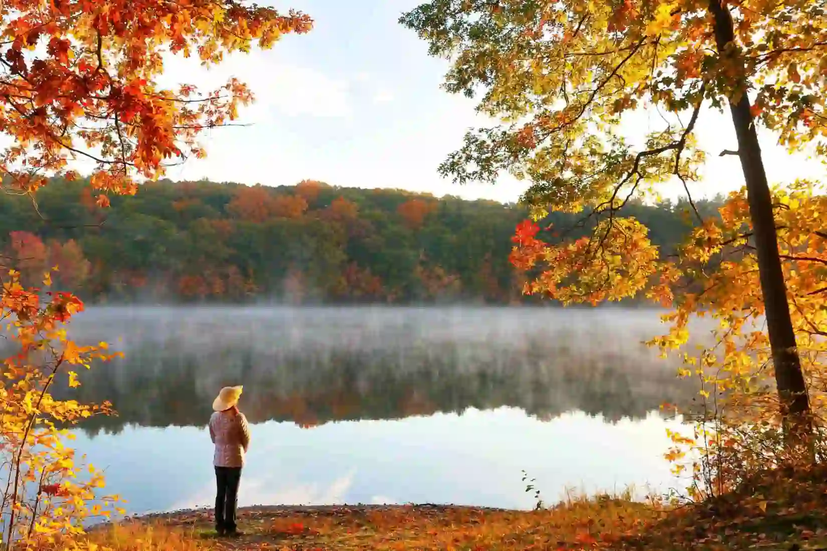 A woman standing by a lake, surrounded by scenic New England autumn foliage with vibrant red, orange, and yellow leaves covering rolling hills