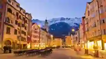 View of a wide road with shops and tall buildings either side. Tables and chairs are laid out in the road and bicycles are parked up on the right side. There is a clock tower which is taller than the rest of the buildings with a turquoise turret on the left, and snowy mountains can be seen at the end of the road, in front of light blue evening sky