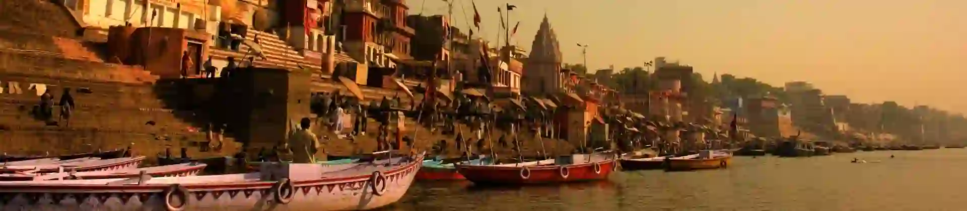 Boats anchored along the ghats of Varanasi at sunrise, with ancient sandstone buildings and temples glowing in golden light by the River Ganges