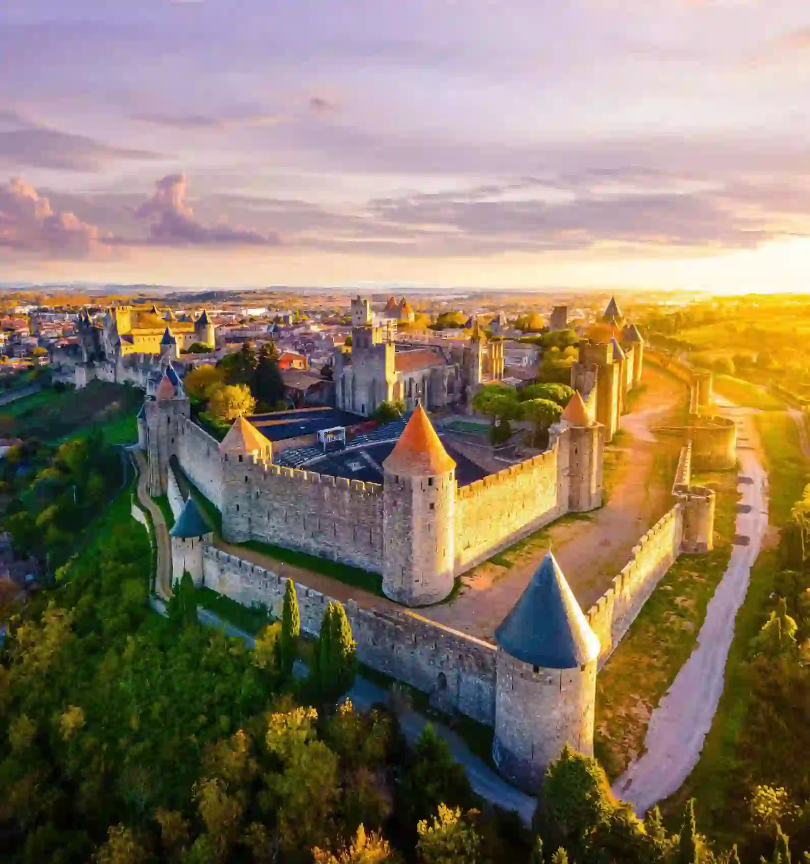 Bird's eye view of the Castle of Carcassonne at sunset, surrounded by trees and grass, and a pathway that loops around the castle. The castle has towers with red and blue turrets, and a distant view of a town can be seen behind it