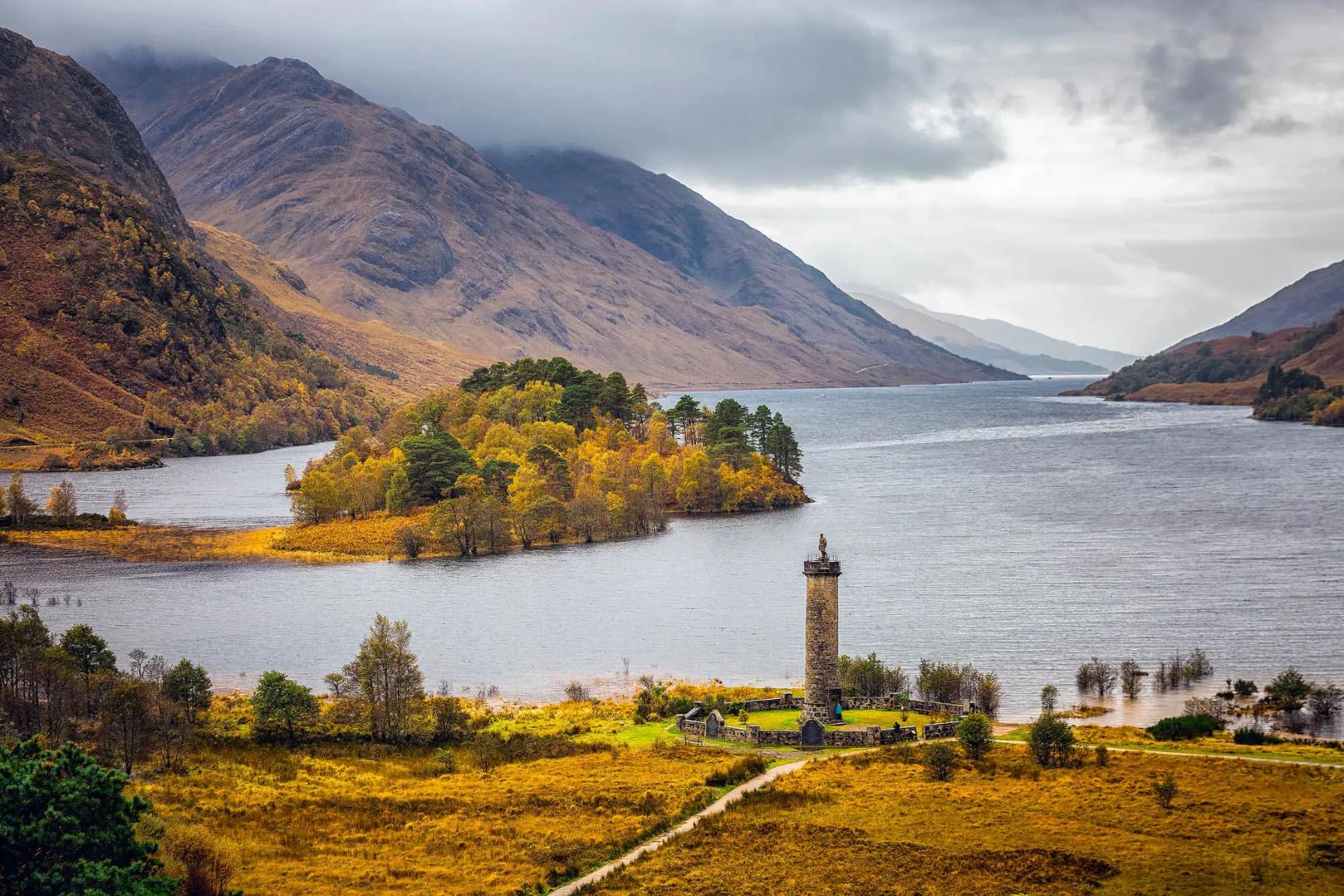 Glenfinnan Monument on a grassy hill at the head of Loch Shiel in the Scottish Highlands, with a lone statue of a Highlander atop the stone tower and dramatic mountains in the background