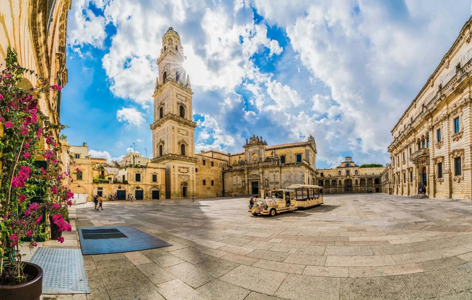 A square in Lecce, Italy, featuring a historic tower and a land train for tourists