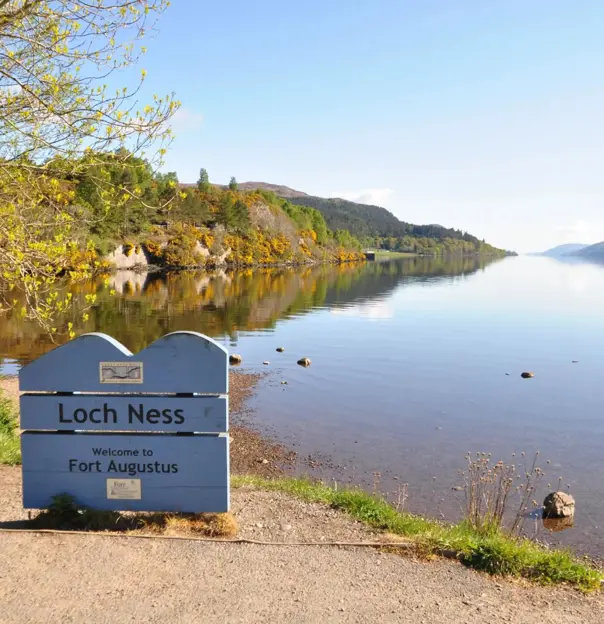 Calm waters of Loch Ness reflecting the surrounding hills under a clear blue sky, with a sign welcoming visitors to Fort Augustus on the shore