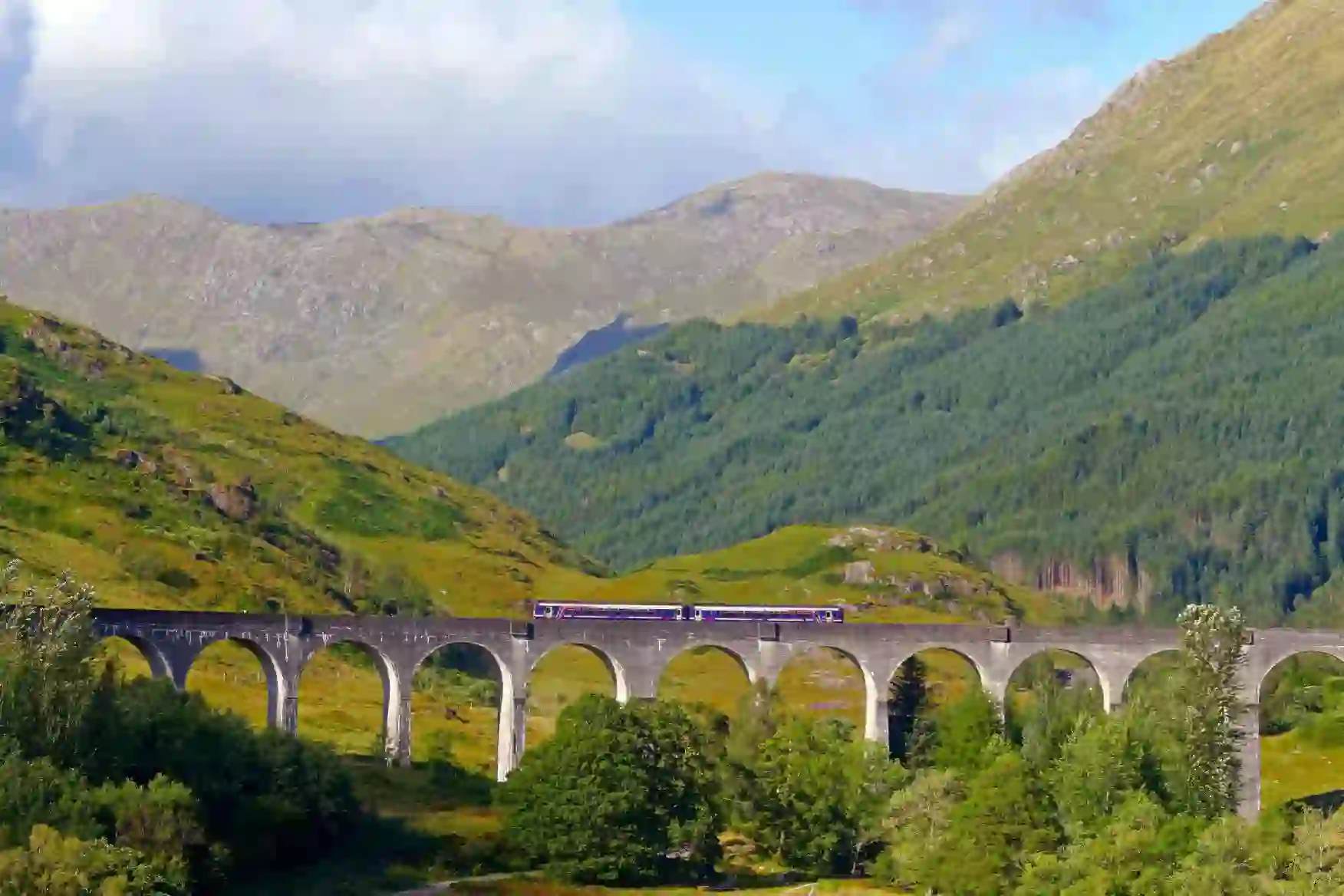 A scenic train travels across the Glenfinnan Viaduct in Scotland, surrounded by rolling green hills and forested mountains