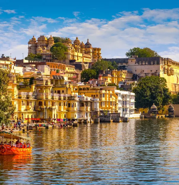 Scenic view of Udaipur’s City Palace and colourful lakeside buildings reflected in Lake Pichola, with a small red boat on the water under a bright blue sky