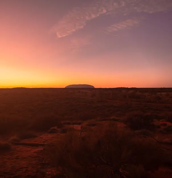 A massive red sandstone monolith rising out of the desert at Uluru in Australia, surrounded by sparse vegetation and open landscape.