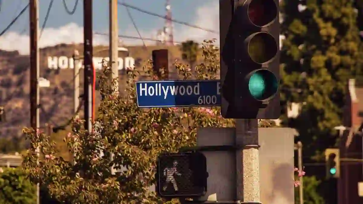A Hollywood Boulevard road sign next to a traffic light and a white pedestrian crossing signal showing a walking man, with the Hollywood Sign visible in the background