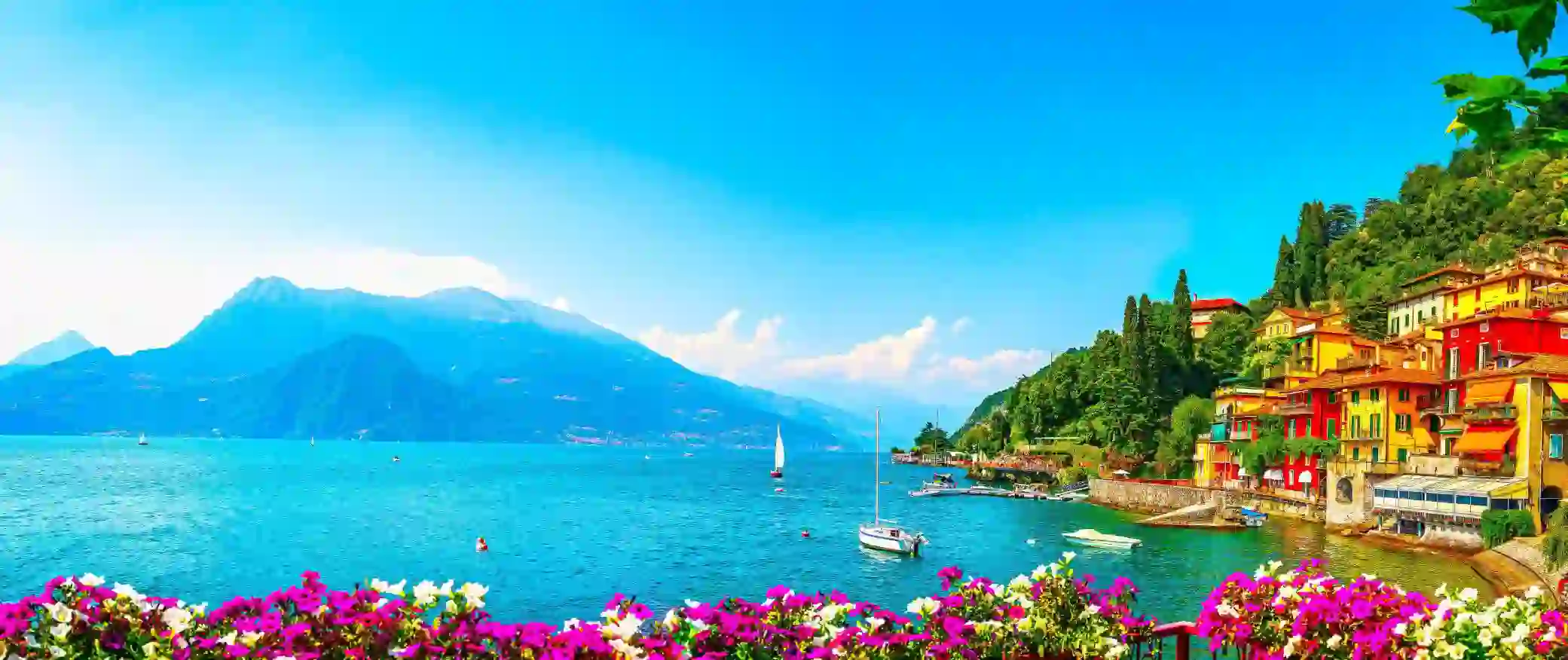 Colourful hillside houses in Lake Como overlooking the lake, where a couple of boats gently float, with mountains in the background